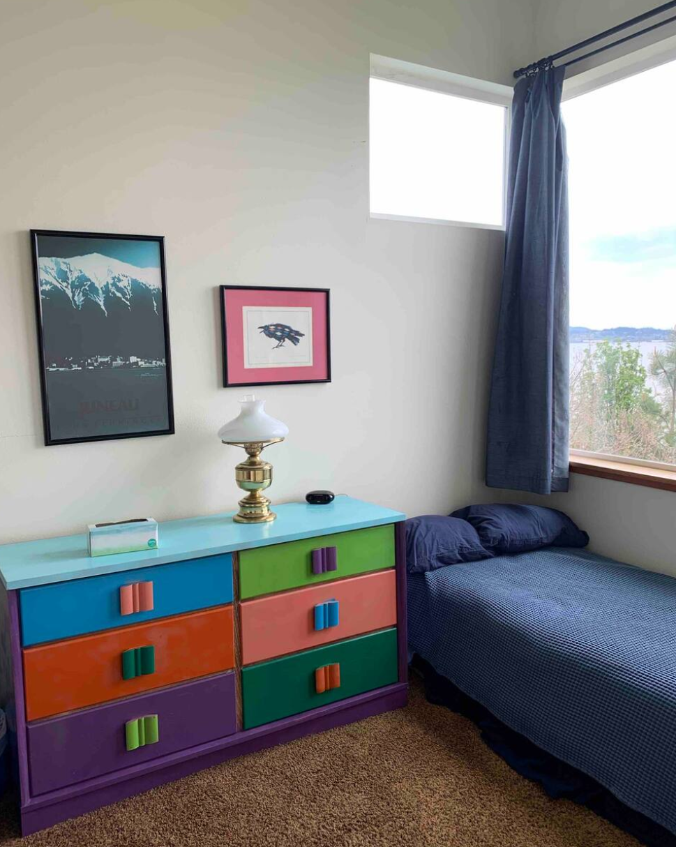 Colorful dresser in a bedroom with a bed and framed pictures on the wall.